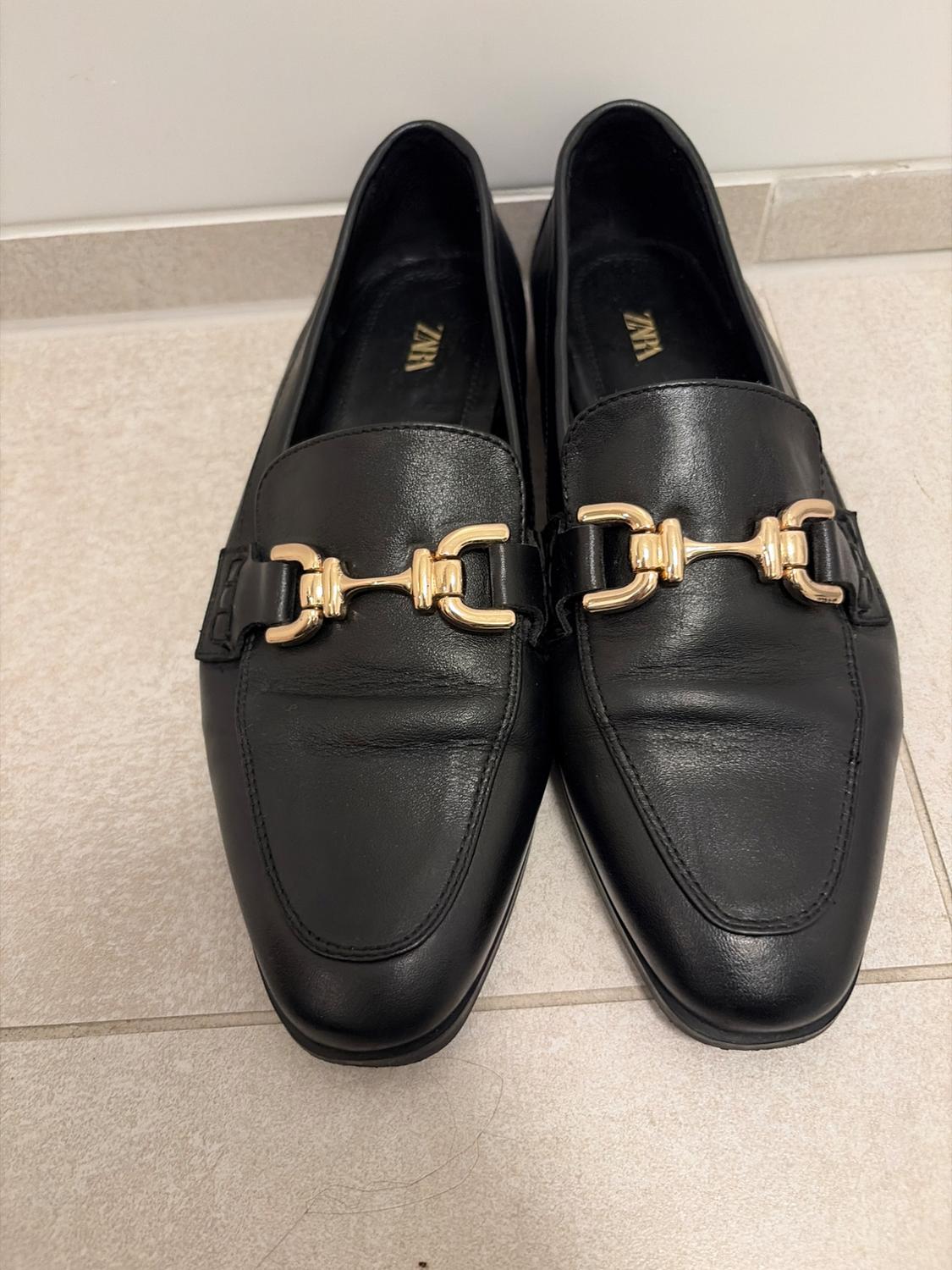 Маύρα mocasines με χρυσή μεταλλική λεπτομέρεια. Στον πάτο αναγράφεται Zara. Βρίσκονται πάνω σε ανοιχτόχρωμο πλακοστρωμένο δάπεδο.