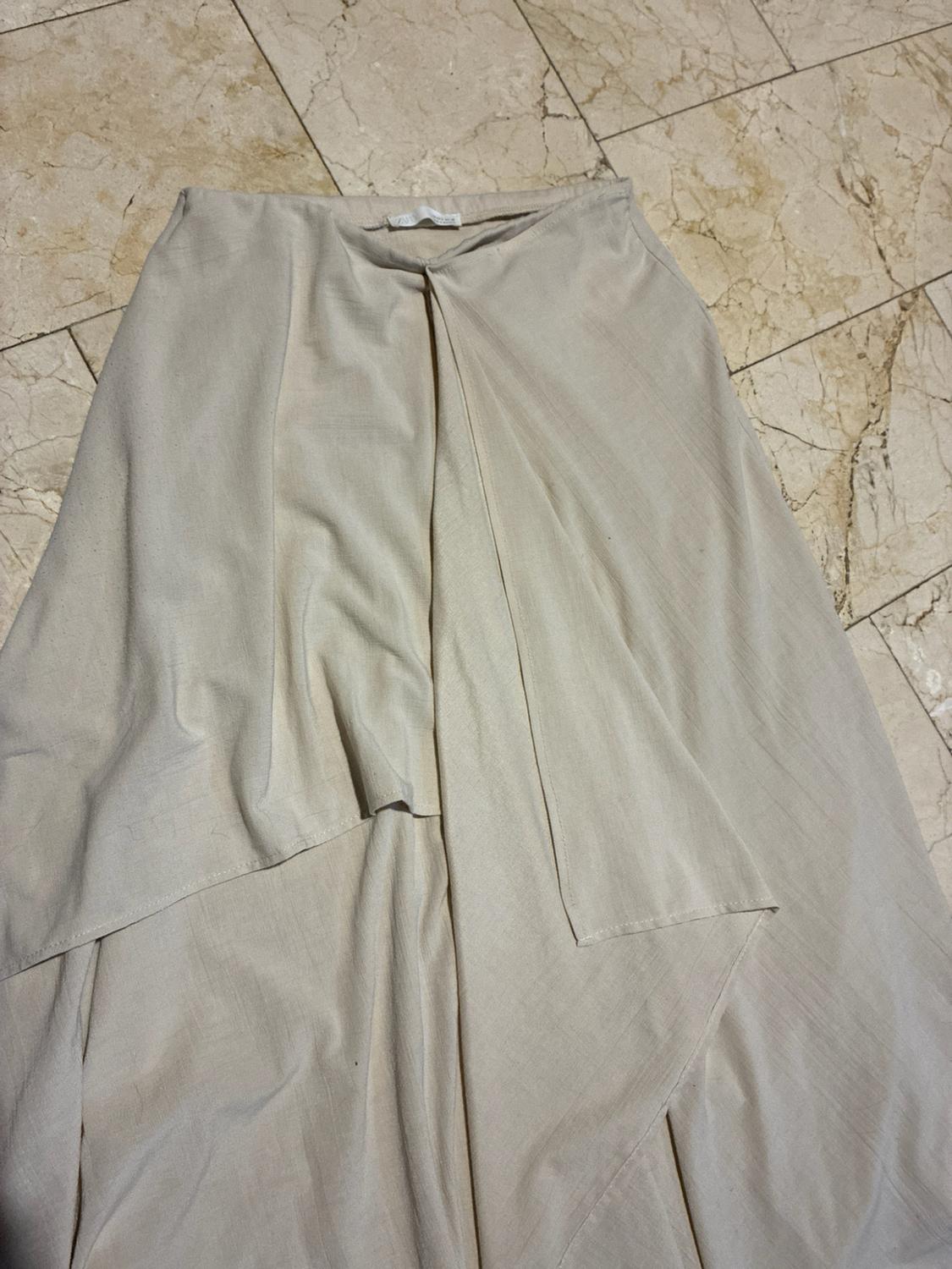 Beige long skirt with an asymmetric design and overlapping panels, spread out on a light tiled floor.