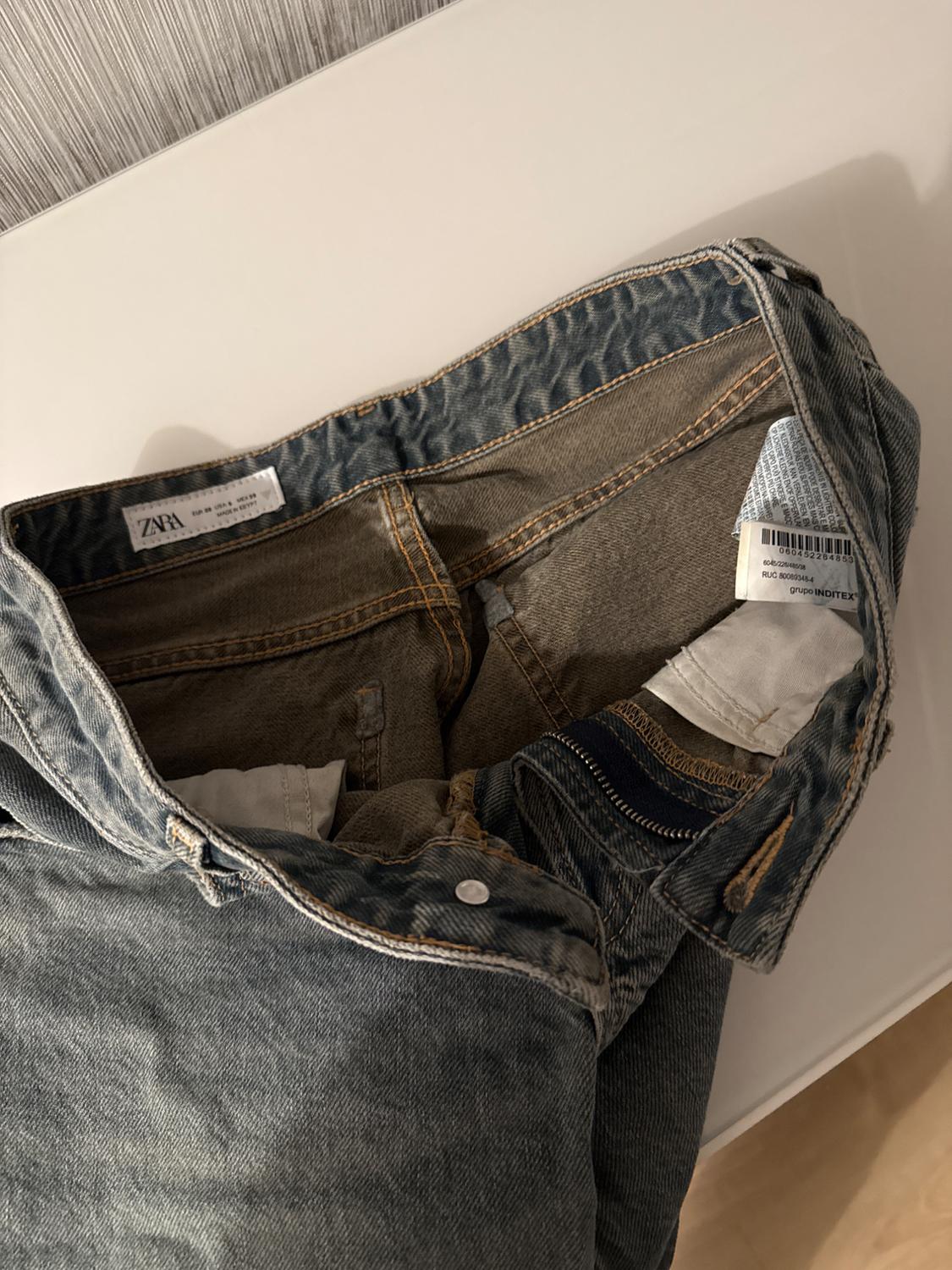 Blue jeans folded on a white surface, displaying inner labels with the "Zara" brand and visible stitching details.