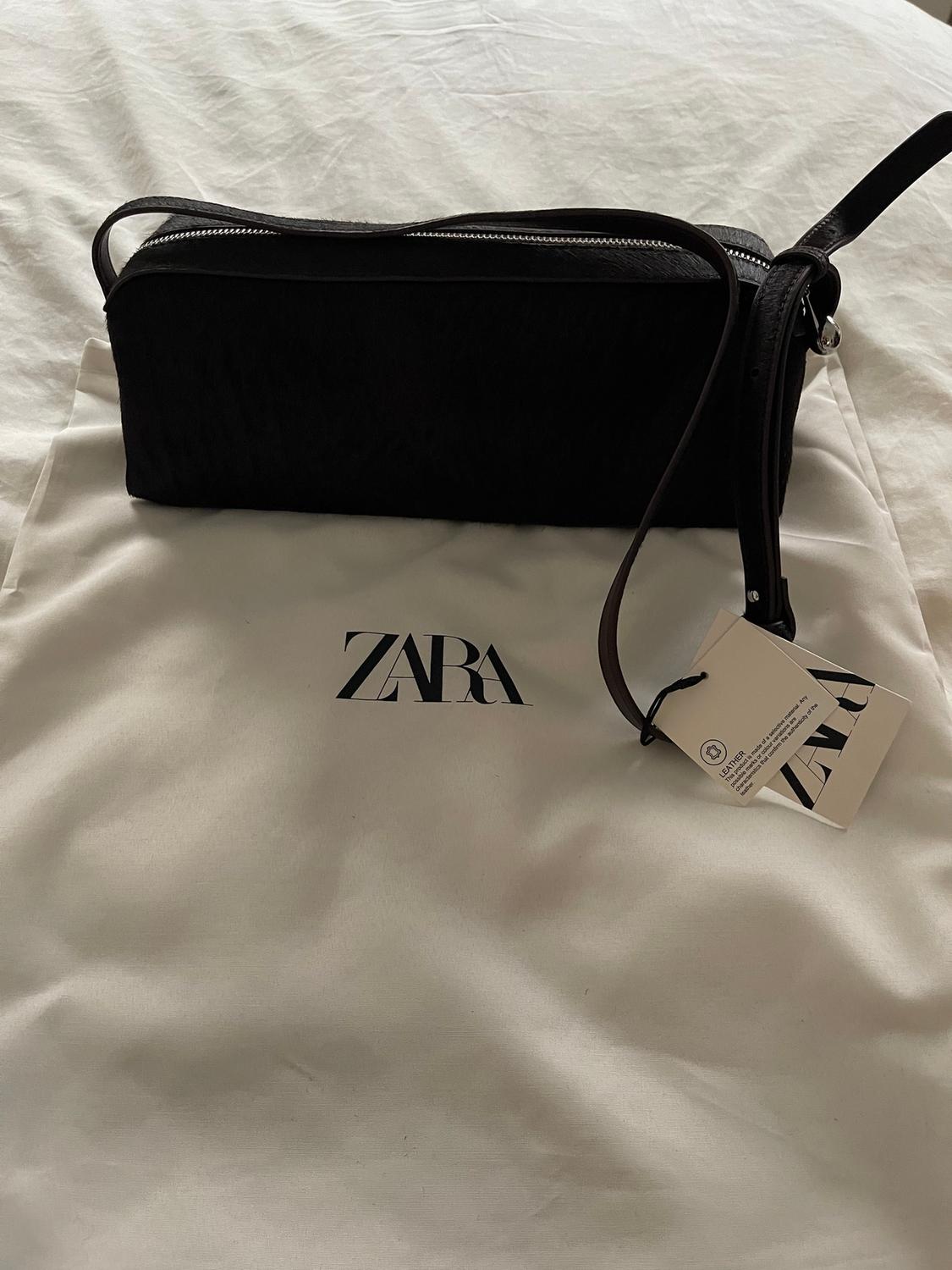 Black handbag with zip and strap on top of a white bag with the "ZARA" logo. Hanging tag visible.
