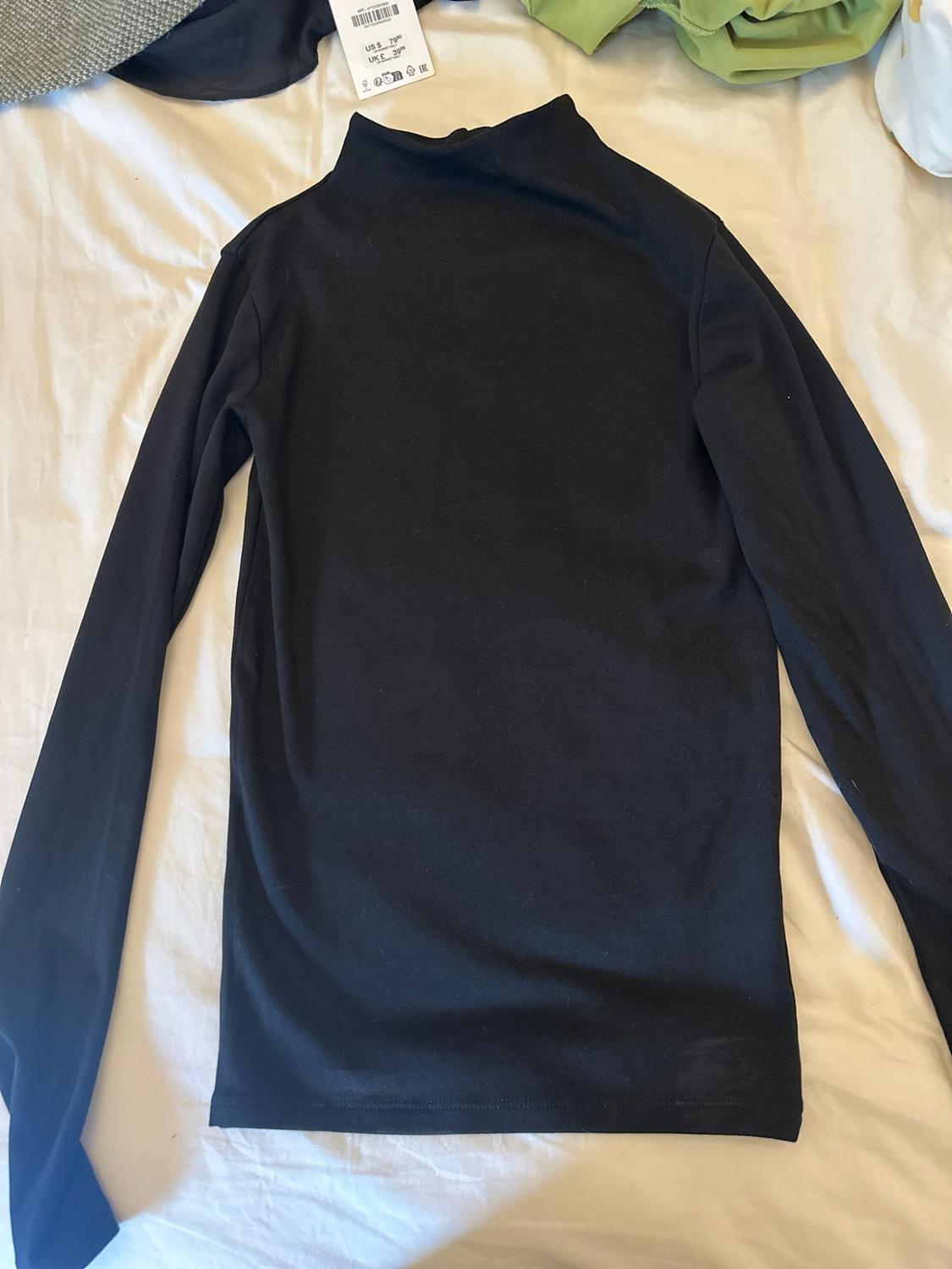 Black long-sleeved, high-necked T-shirt laid out on a white sheet. Other garments and a label can be seen at the top.