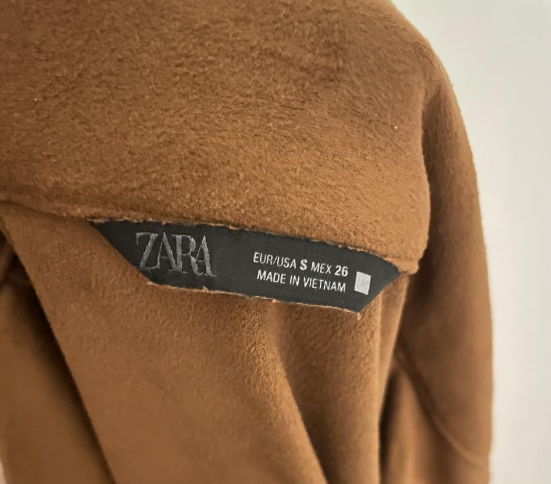 Μαύρη ετικέτα σε καφέ ένδυμα με κείμενο: "ZARA EUR/USA S MEX 26 MADE IN VIETNAM". Απαλό ύφασμα και ορατή υφή.