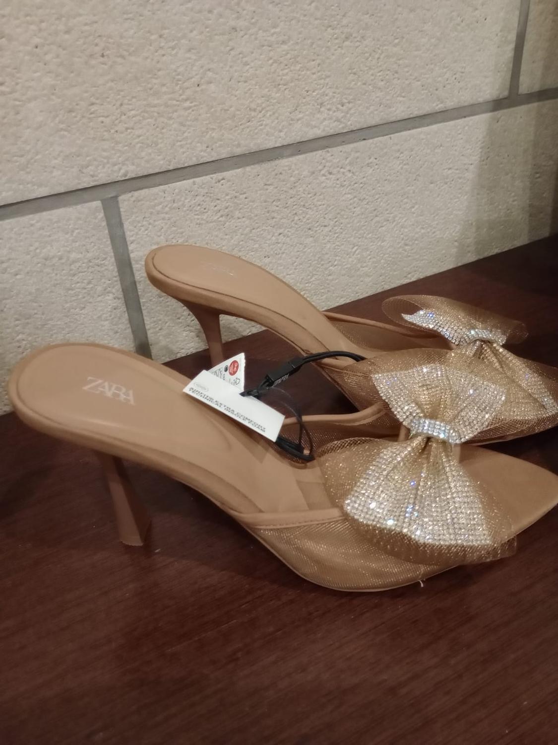 Beige high heels with decorative sparkly bow, visible label with Zara logo, beige wall background and wooden surface.