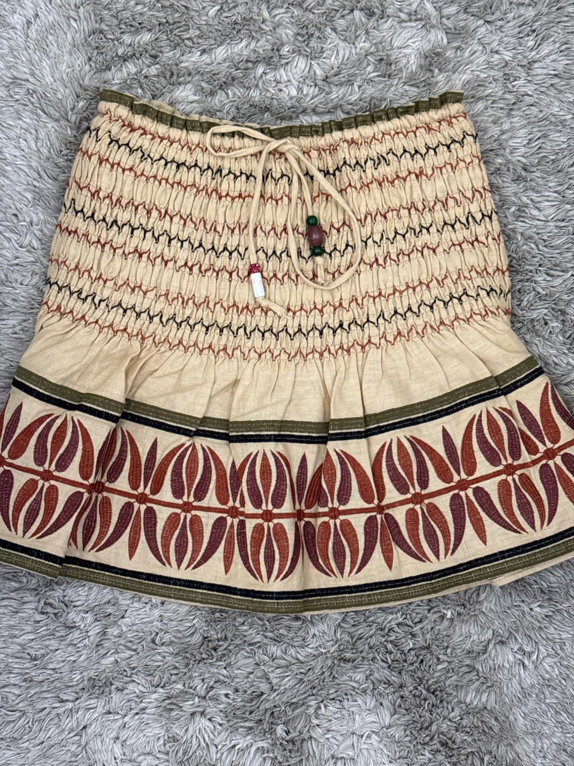 Short beige skirt on a grey shag pile rug. Gathered waist with drawstring and beads, hem with reddish leaf motifs and green lines.