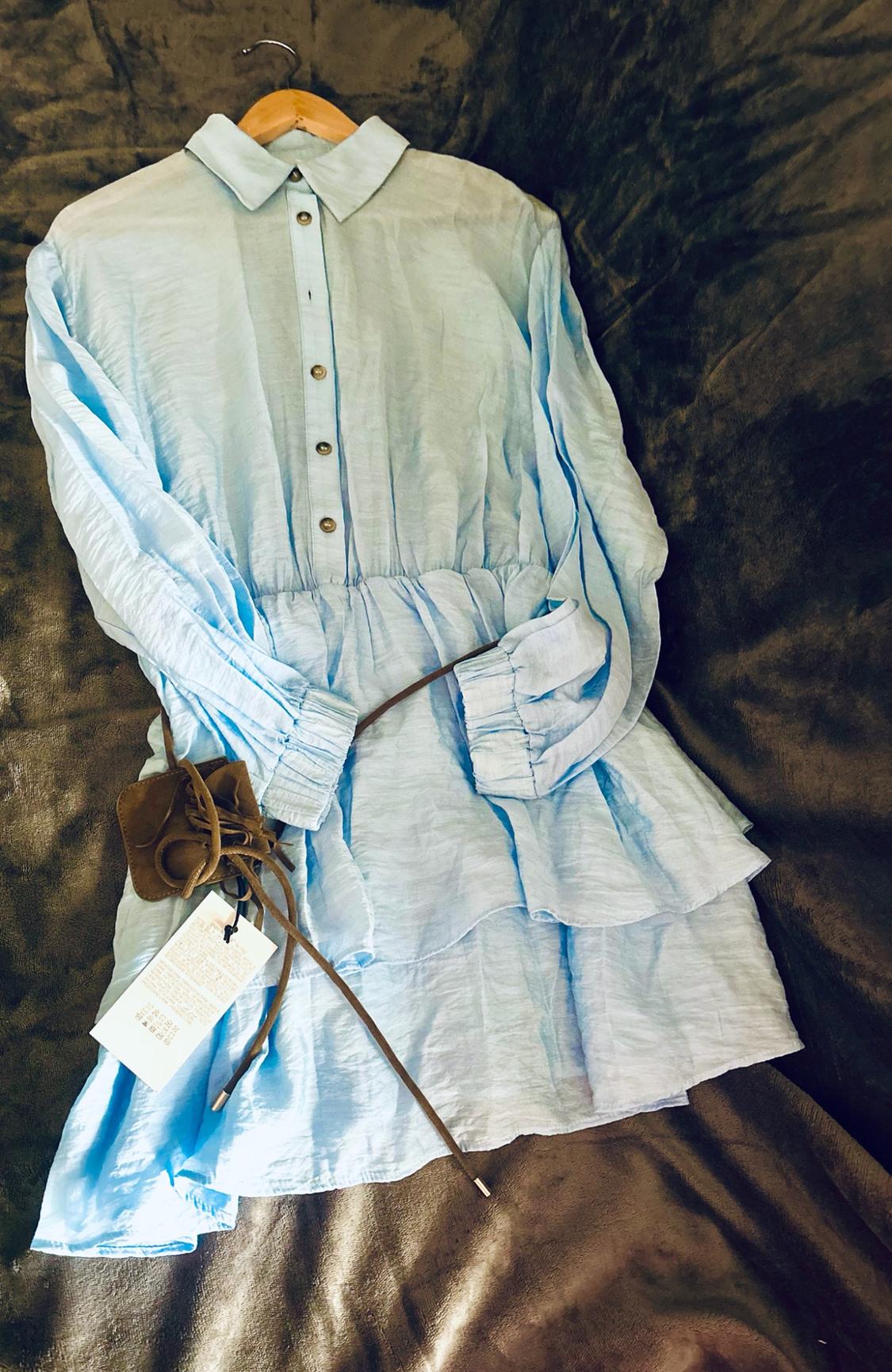 Γαλάζιο φόρεμα με κουμπιά, βολάν και μακριά μανίκια, κρεμασμένο σε ξύνηστρα σε σκούρο φόντο. Διαθέτει ένα καφέ αξεσουάρ με ετικέτα.