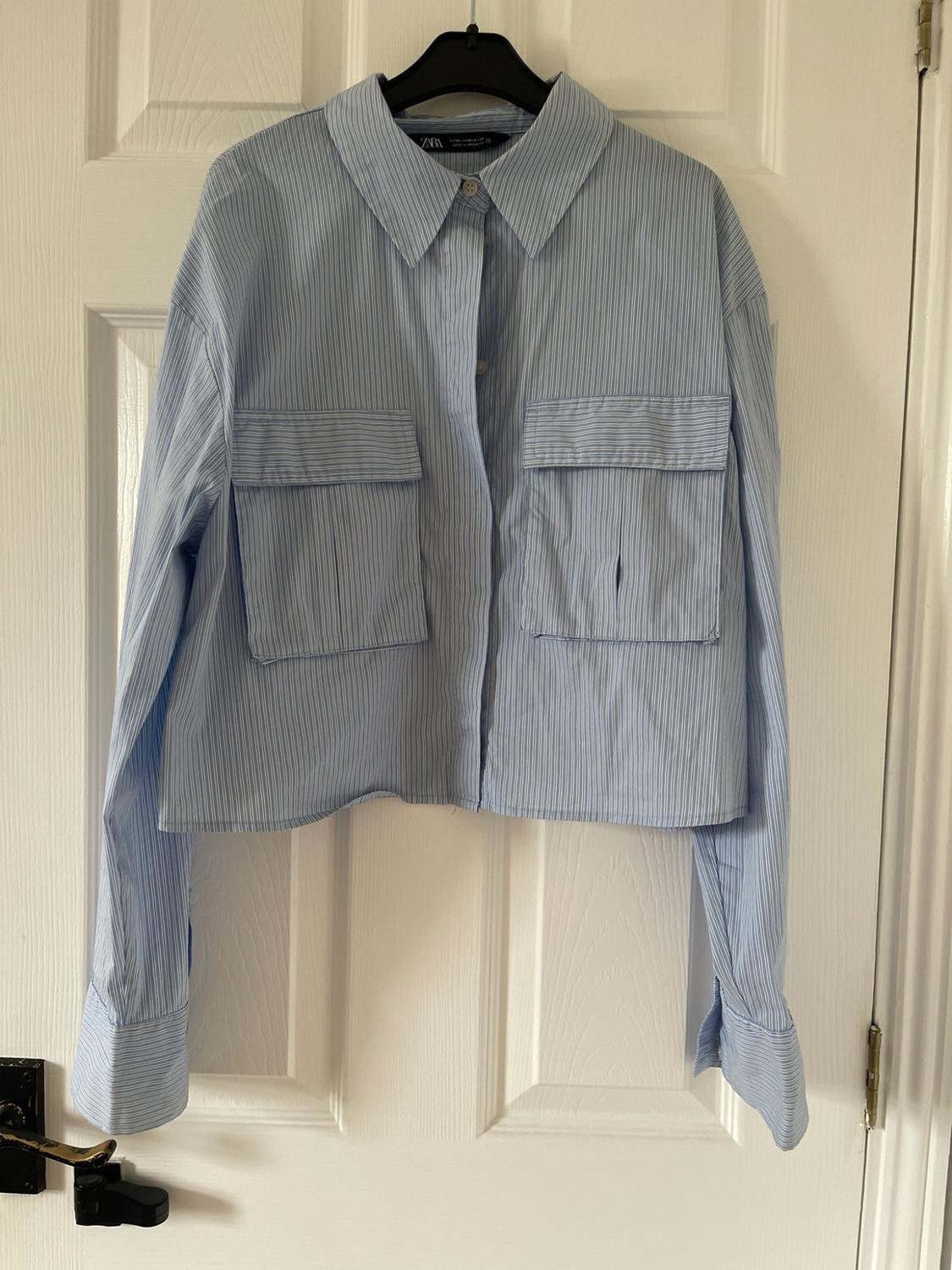 Cropped long-sleeved shirt with blue and white stripes and chest pockets, hanging on a white door. Label with ZARA text.