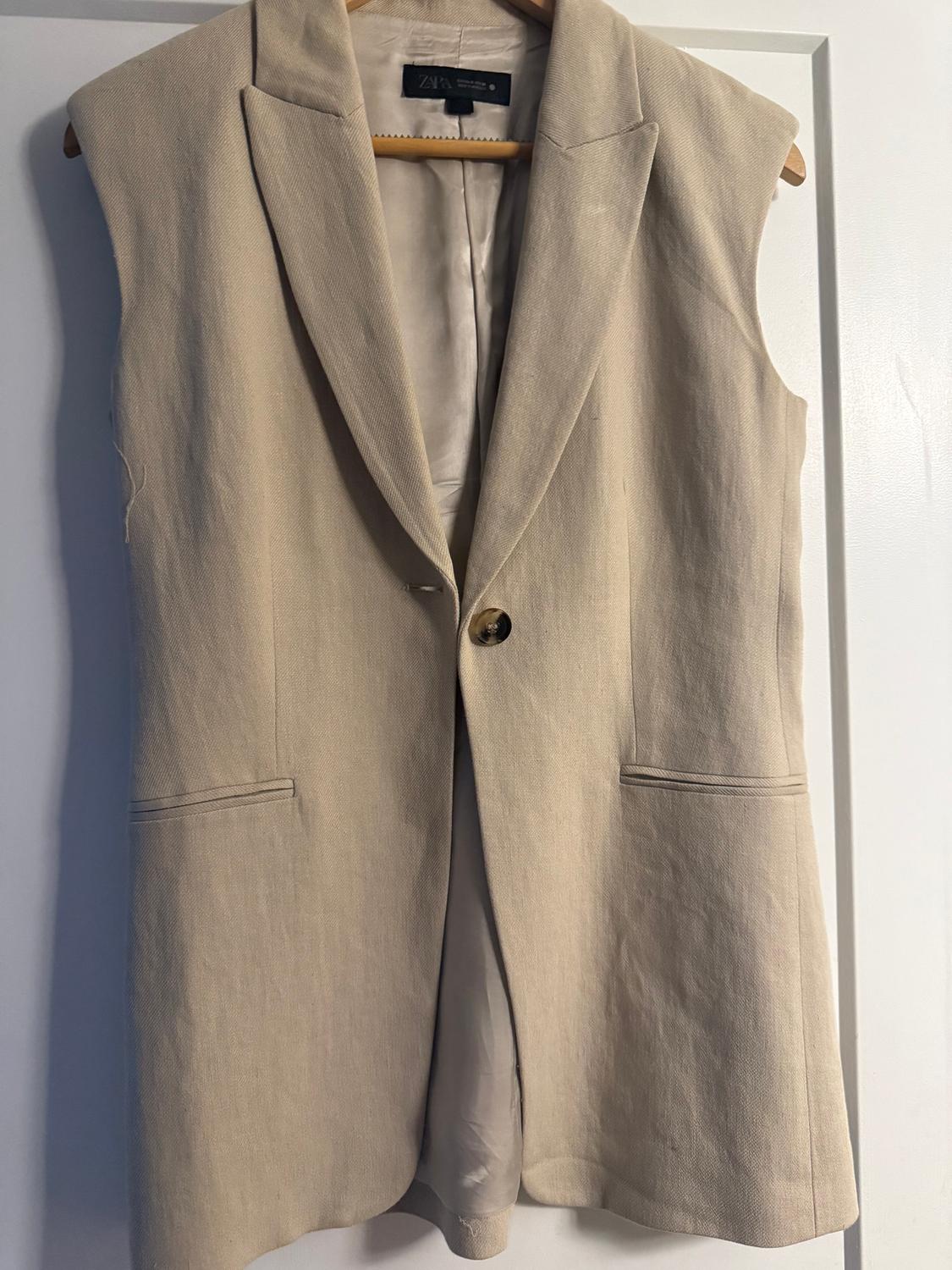 Beige sleeveless waistcoat hanging on a wooden hanger, with lapels and a front button. White background.