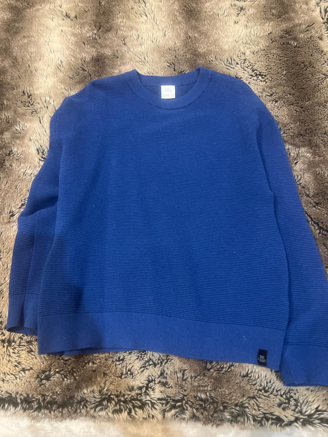 Fine-knit blue jumper on a shaggy beige and brown rug. Visible label on the collar with the text "ZARA".