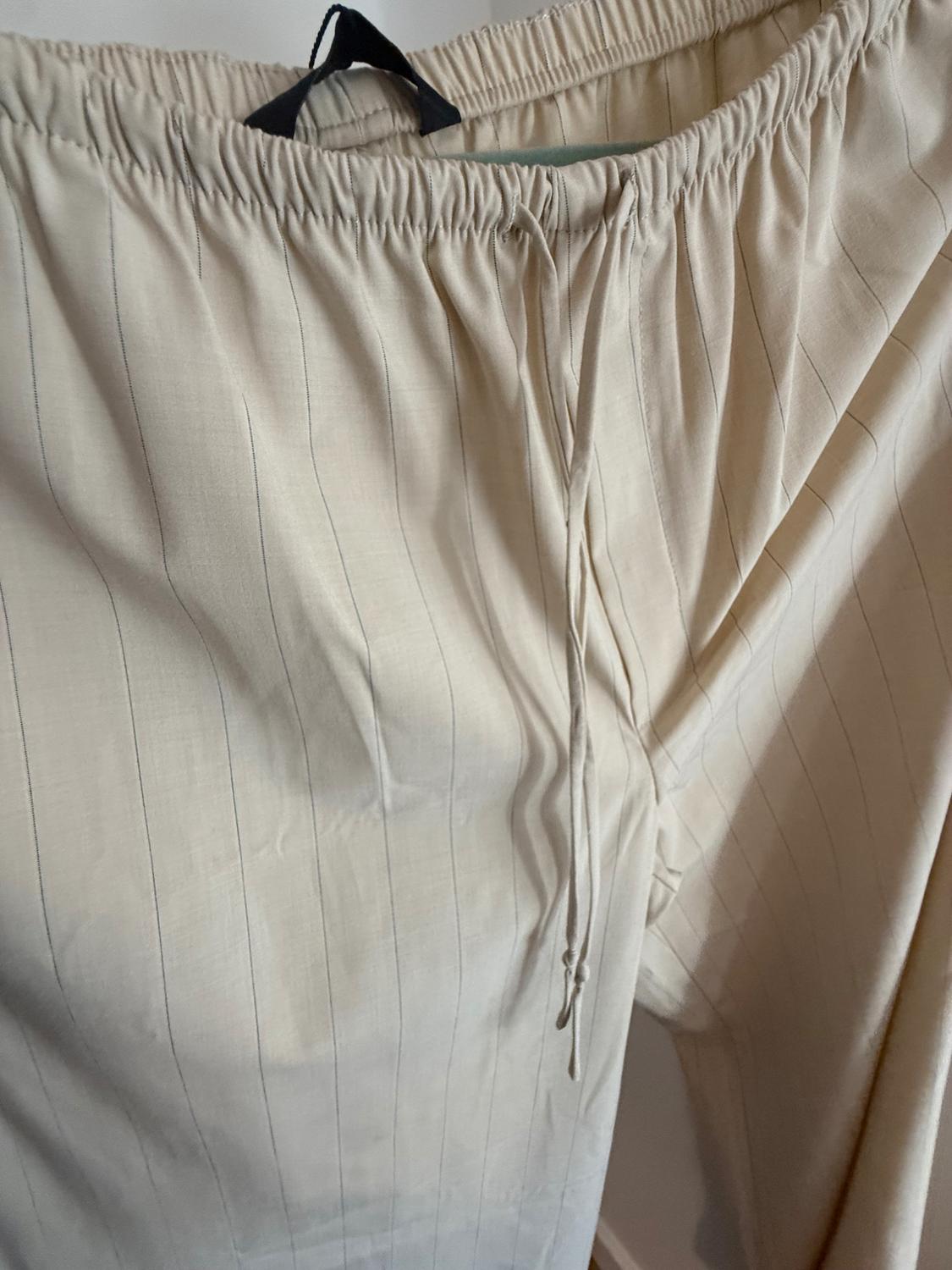 Close-up of beige trousers with fine dark vertical stripes. Highlights include a gathered elasticated waistband with an adjustable drawstring and a black label.