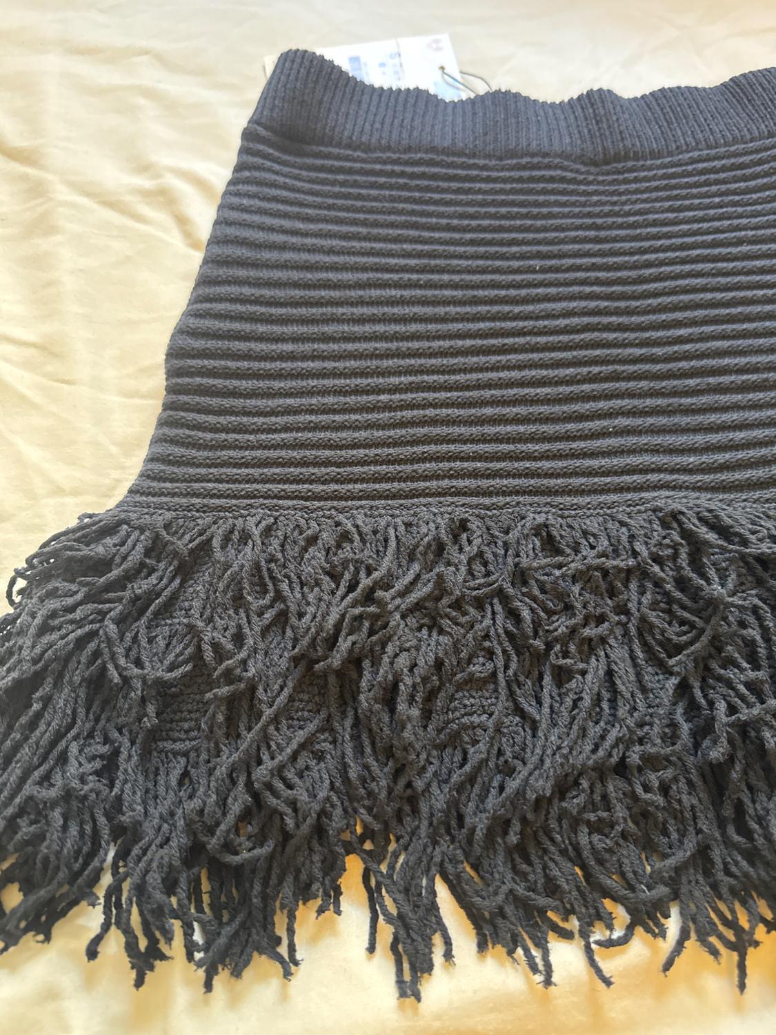 Close-up of a black ribbed knit skirt with a long fringed hem, laid out on a light beige fabric surface.