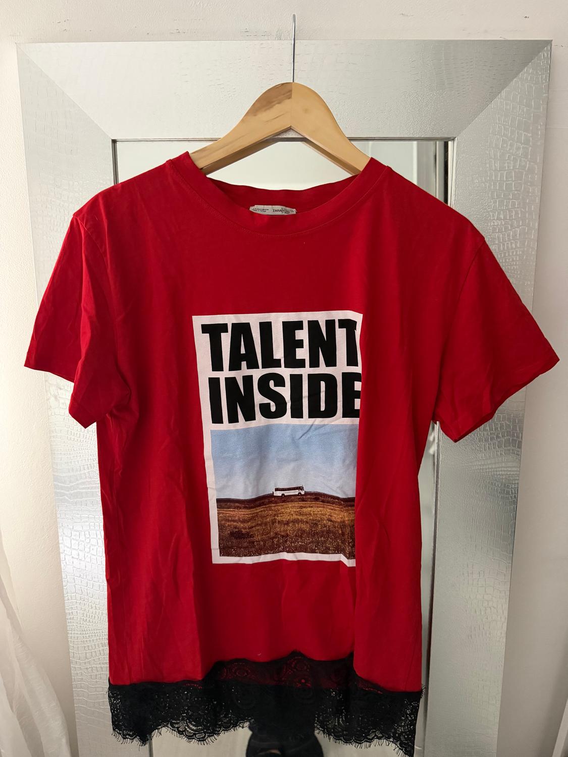 Red T-shirt with "TALENT INSIDE" text and a landscape in the centre. Black lace detail on the bottom hem. Hung on a hanger.
