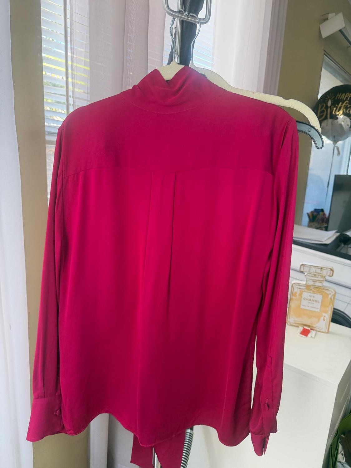Red blouse hanging on a hanger in front of a window with white curtains. In the background, a perfume bottle on a piece of furniture.