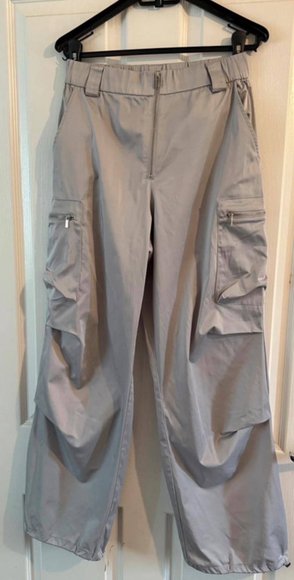 Grey cargo trousers hanging on a black hanger, with side pockets and a zip closure, over a white door.