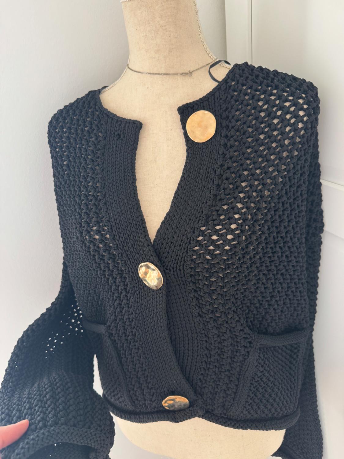 Mannequin wearing a black open-knit cardigan with a V-neck, front pockets, and large gold buttons against a light background.