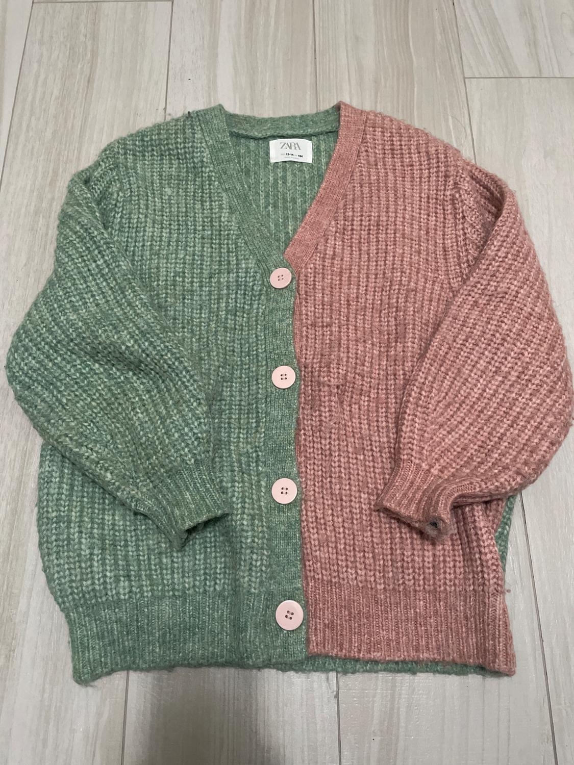 A two-tone knitted cardigan, green and pink, with pink buttons, placed on a light wooden floor. The visible label reads: "ZARA".