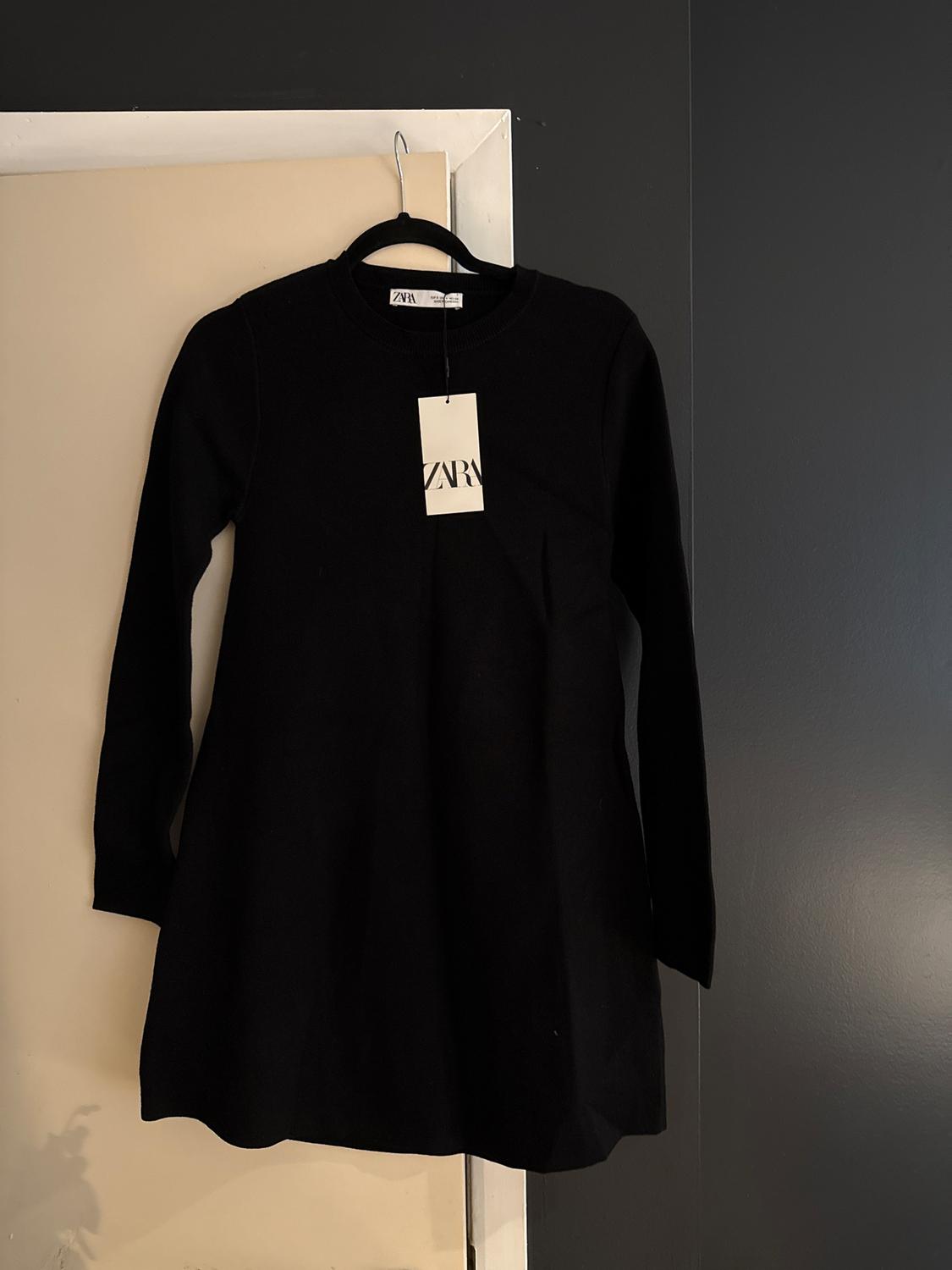 Black long-sleeved dress hanging on a hanger with a Zara tag, over a white door and dark wall.