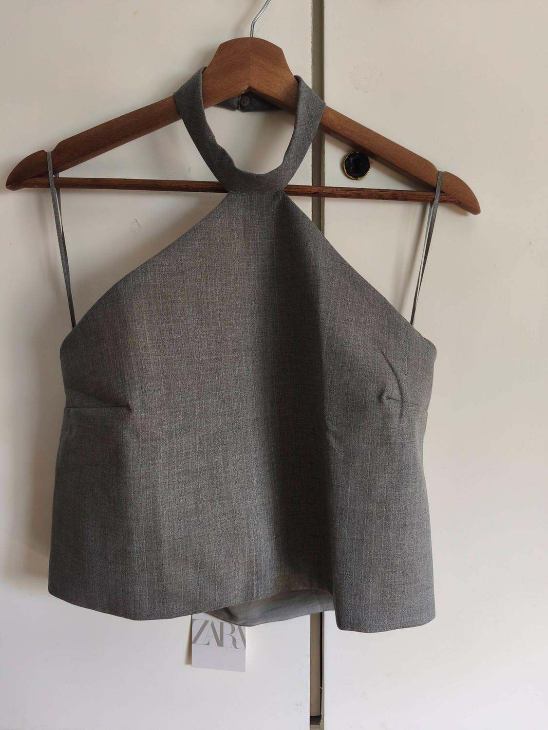 Grey sleeveless halter neck top on a wooden hanger. Visible "ZARA" tag. White background.