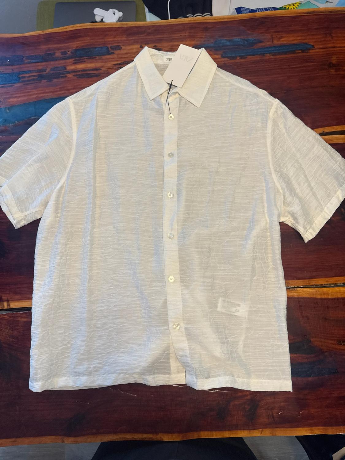 Short-sleeve textured white shirt with front buttons, laid out on a dark wooden table. ZARA label on the collar.