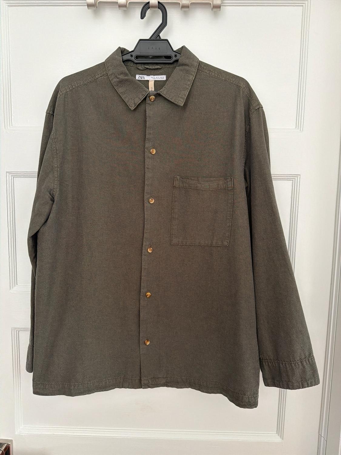 Long-sleeve green button-down shirt with a pocket, hanging on a hanger over a white door. Label with the text ZARA.