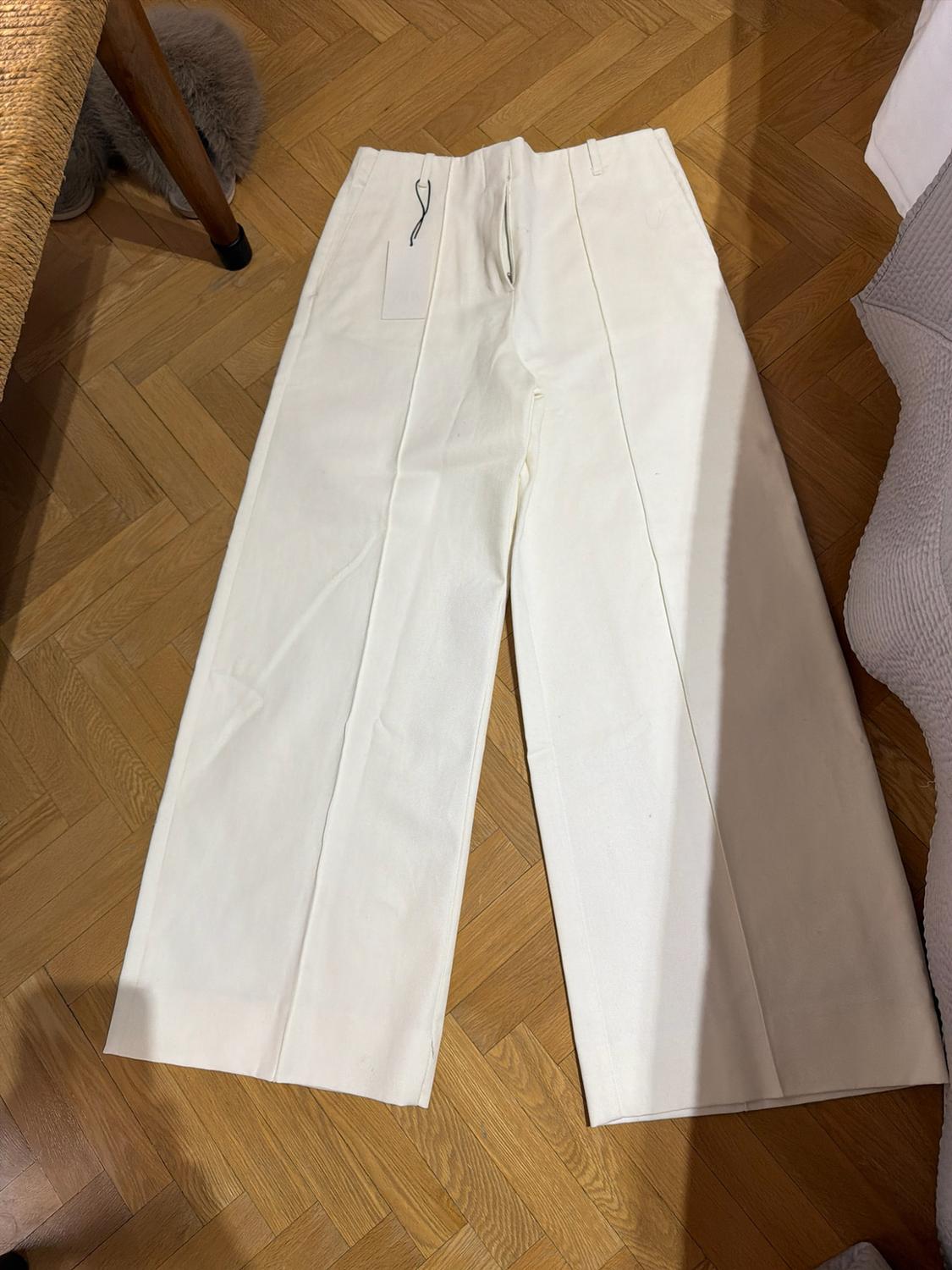 White wide-leg trousers with front seam and label, laid out on a herringbone wooden floor in a relaxed setting.