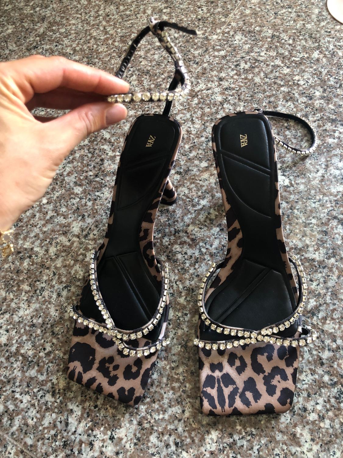 Hand holding the strap of a pair of leopard-print, embellished high-heeled sandals on a granite floor. ZARA text on the insole.