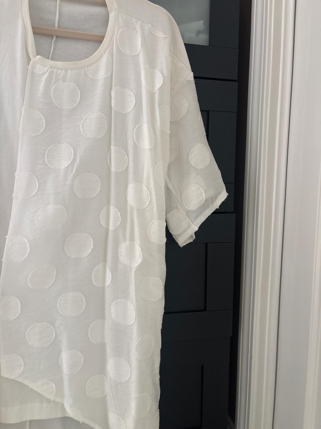 White t-shirt with embossed polka dot print, hanging on a wooden hanger in front of a dark door.