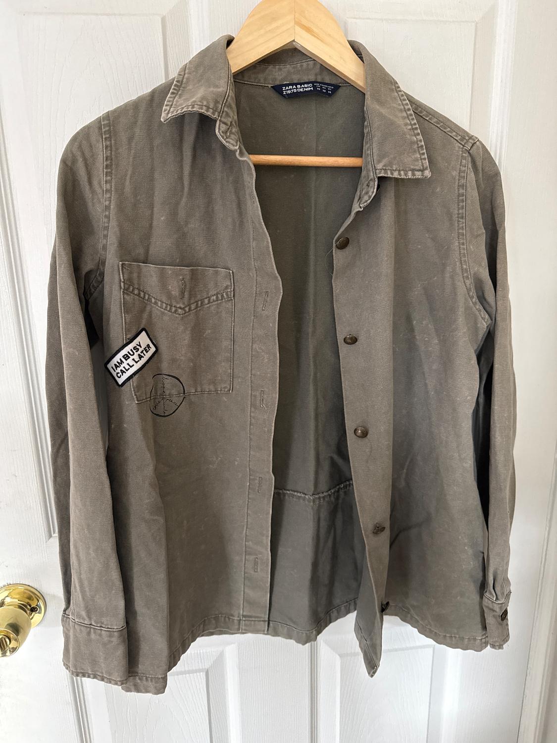 Olive green shirt hanging on a wooden hanger, with a patch on the pocket that says "I AM BUSY CALL LATER". White door in the background.