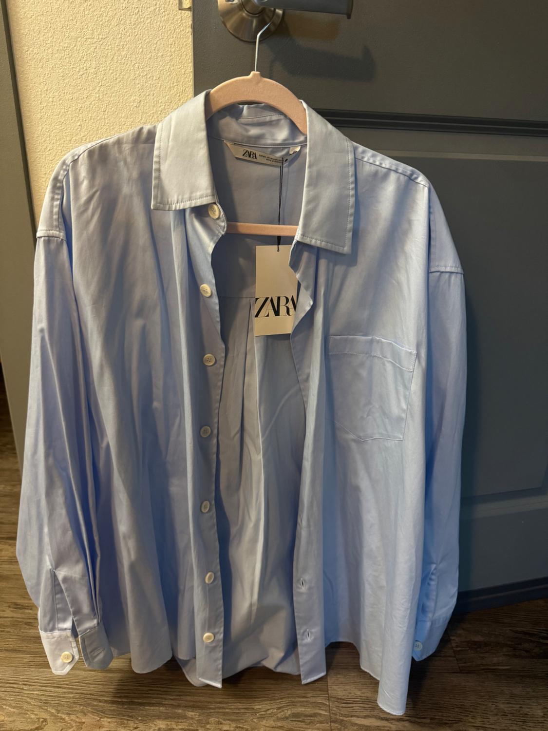 Light blue button-down shirt hanging on a wooden coat hanger, with a visible Zara brand label, against a grey door background.