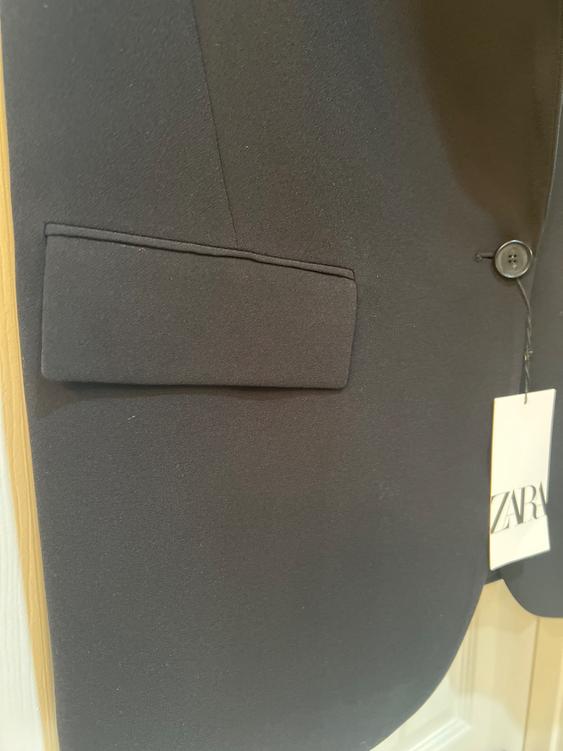 Detail of a black blazer with a pocket flap and visible button. Hanging tag with the text "ZARA" on the right edge.