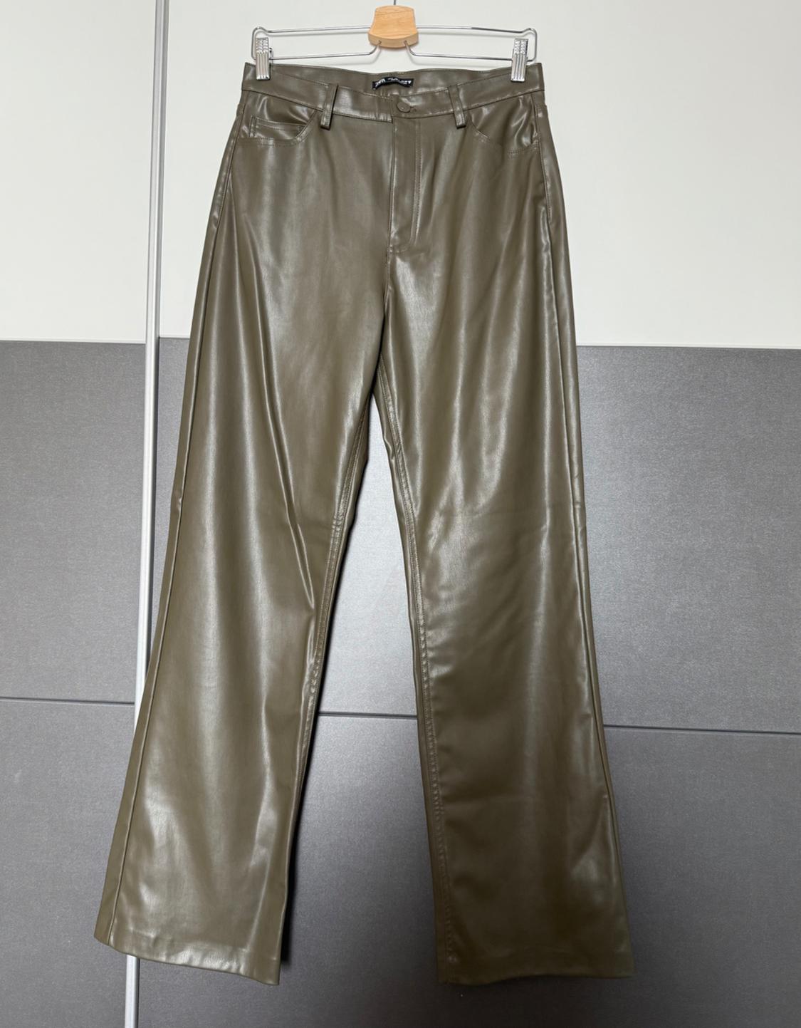 Olive green faux leather trousers hanging on a coat hanger against a grey and white backdrop.