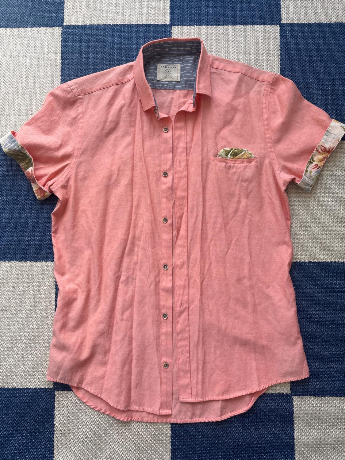 Pink short-sleeved shirt with buttons, pocket, and floral details on the sleeves. Blue and white checkered background. Visible tag.