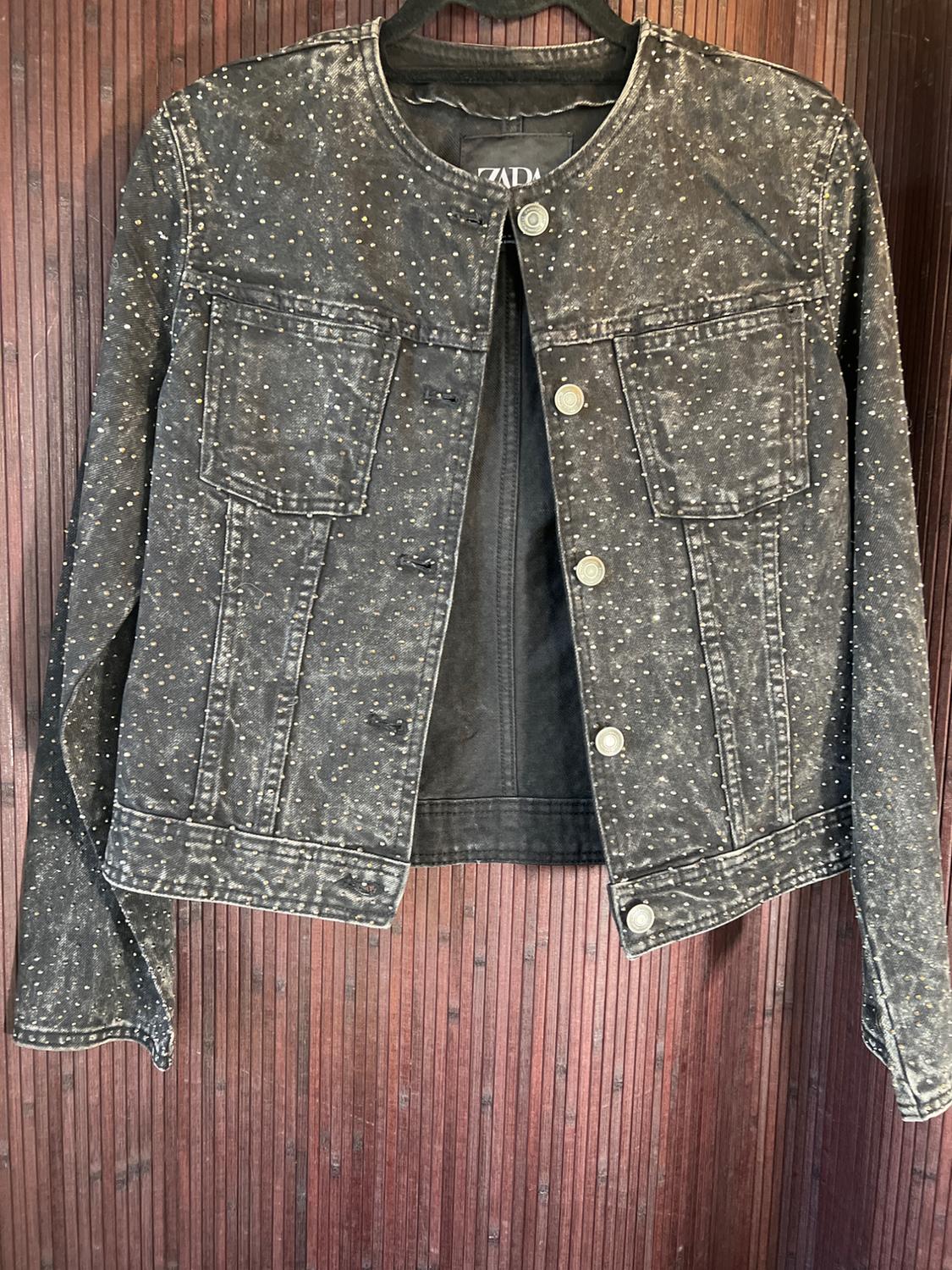 Black jacket with silver studs and metal buttons, hanging on a wooden wall. Visible tag with text "ZARA".