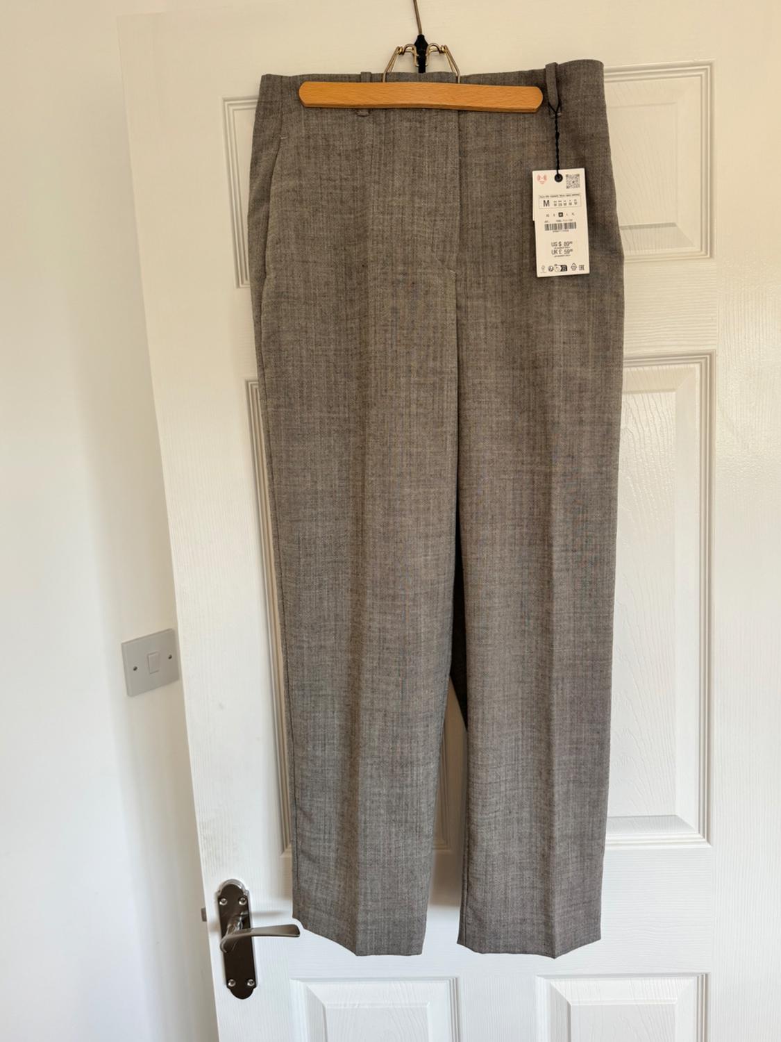 Light grey trousers made of lightweight fabric hanging on a wooden hanger over a white door, with a visible label indicating size M and product details.