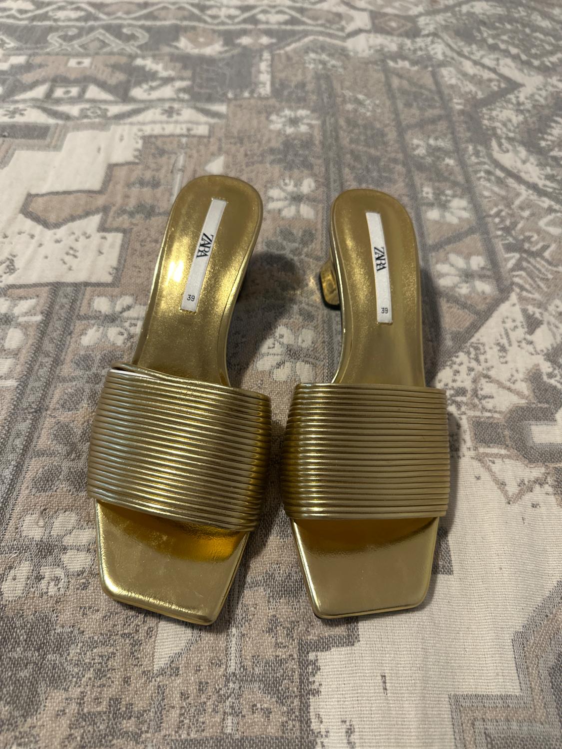 Gold sandals with wide straps and a ribbed texture on a surface with a geometric pattern. Visible label: "ZARA 39".