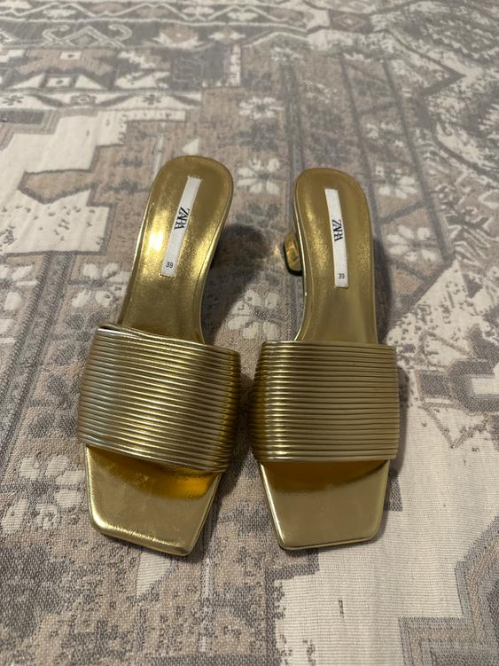 Golden sandals with wide straps and ribbed texture on a surface with a geometric pattern. Visible label: "ZARA 39".