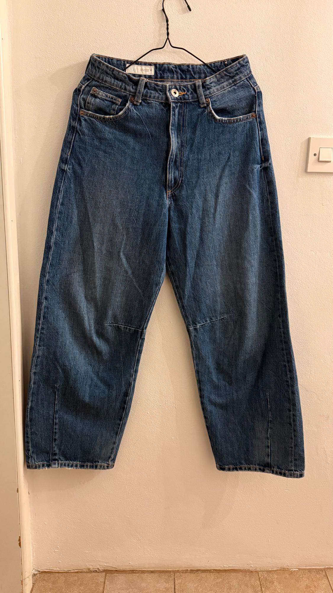 Blue denim jeans hanging on a hanger against a white wall. Loose fit and five visible pockets.