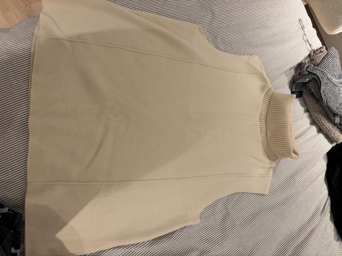 Beige knitted gilet with a ribbed high neck, on a bed with grey and white striped sheets. Folded clothes next to it.