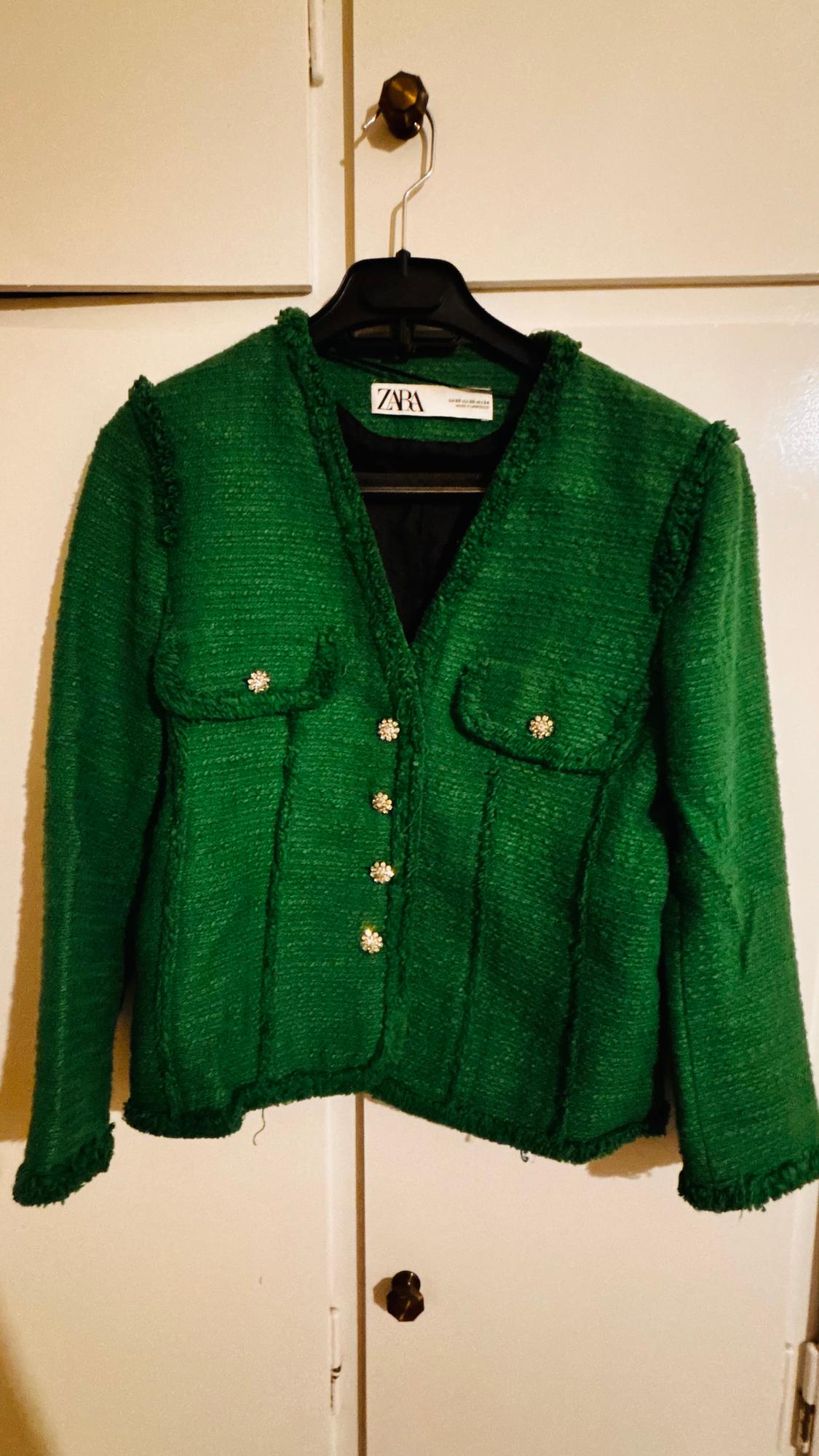 Green Zara jacket hanging on a black hanger, with shiny decorative buttons and front pockets. White wardrobe background.