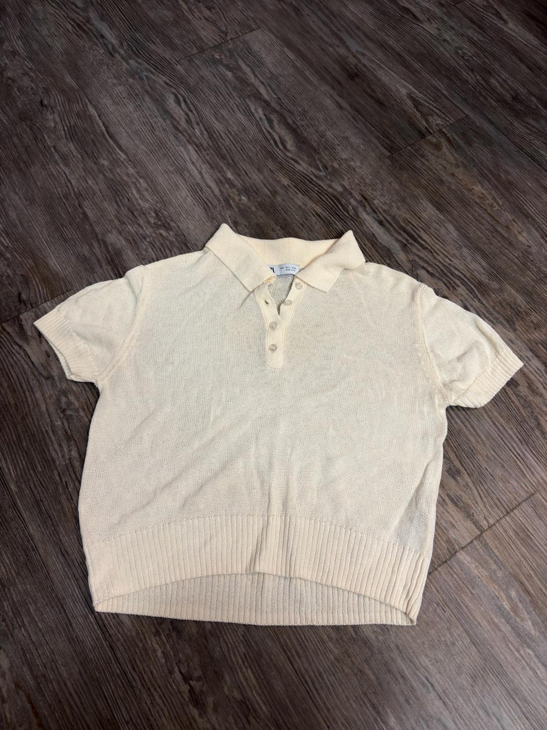 Cream-coloured short-sleeved knitted polo shirt with a collar, buttons and ribbed hem, on a dark wooden floor.