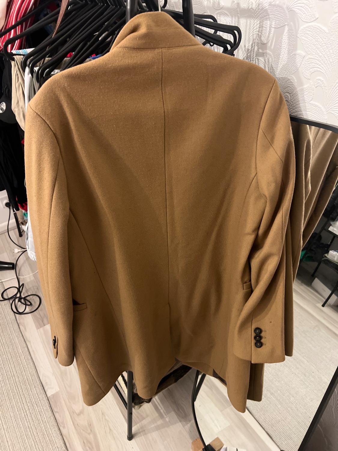 Brown jacket hanging on a coat rack, seen from behind. Background with textured wall and other garments hanging.