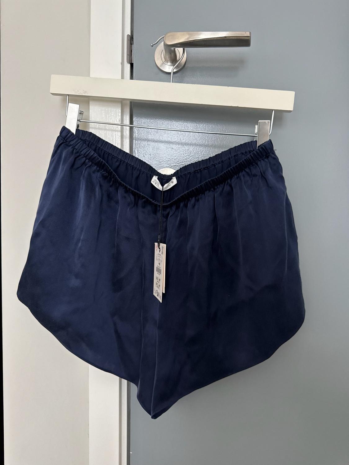 Navy blue satin shorts with an elasticated waist, hanging from a white hanger on the knob of a grey door. ZARA label.