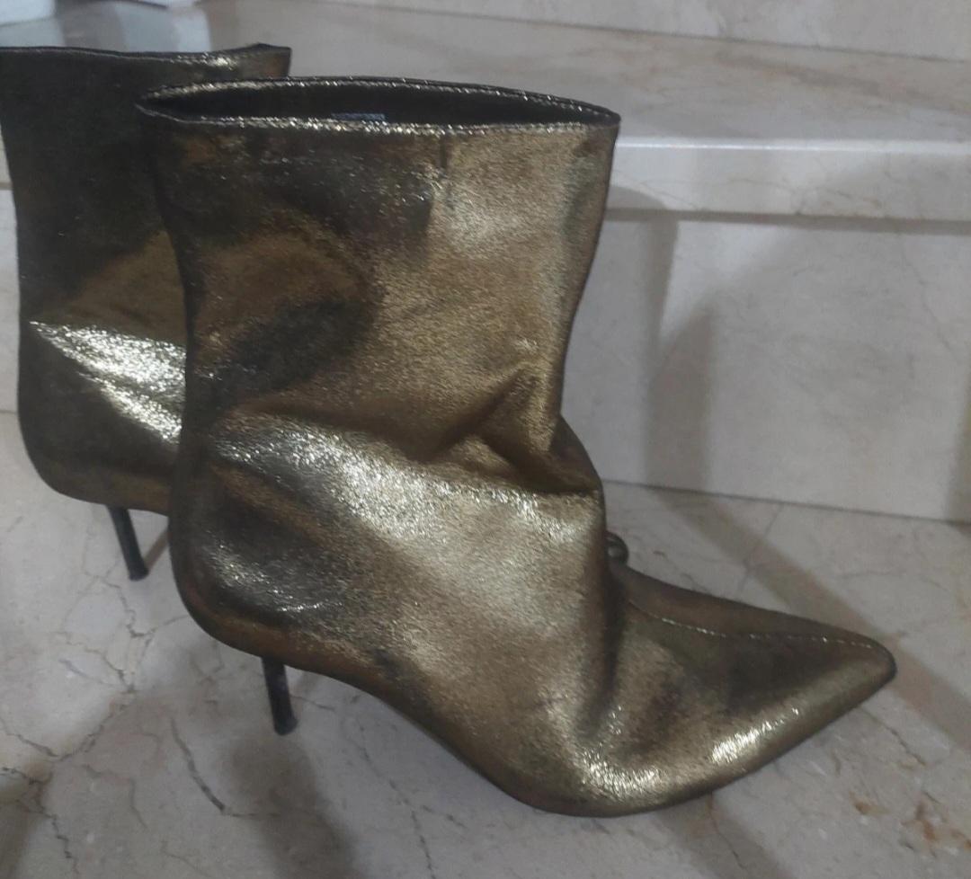Metallic gold ankle boots with a crinkled finish, pointed toe and stiletto heel, resting on light marble steps.