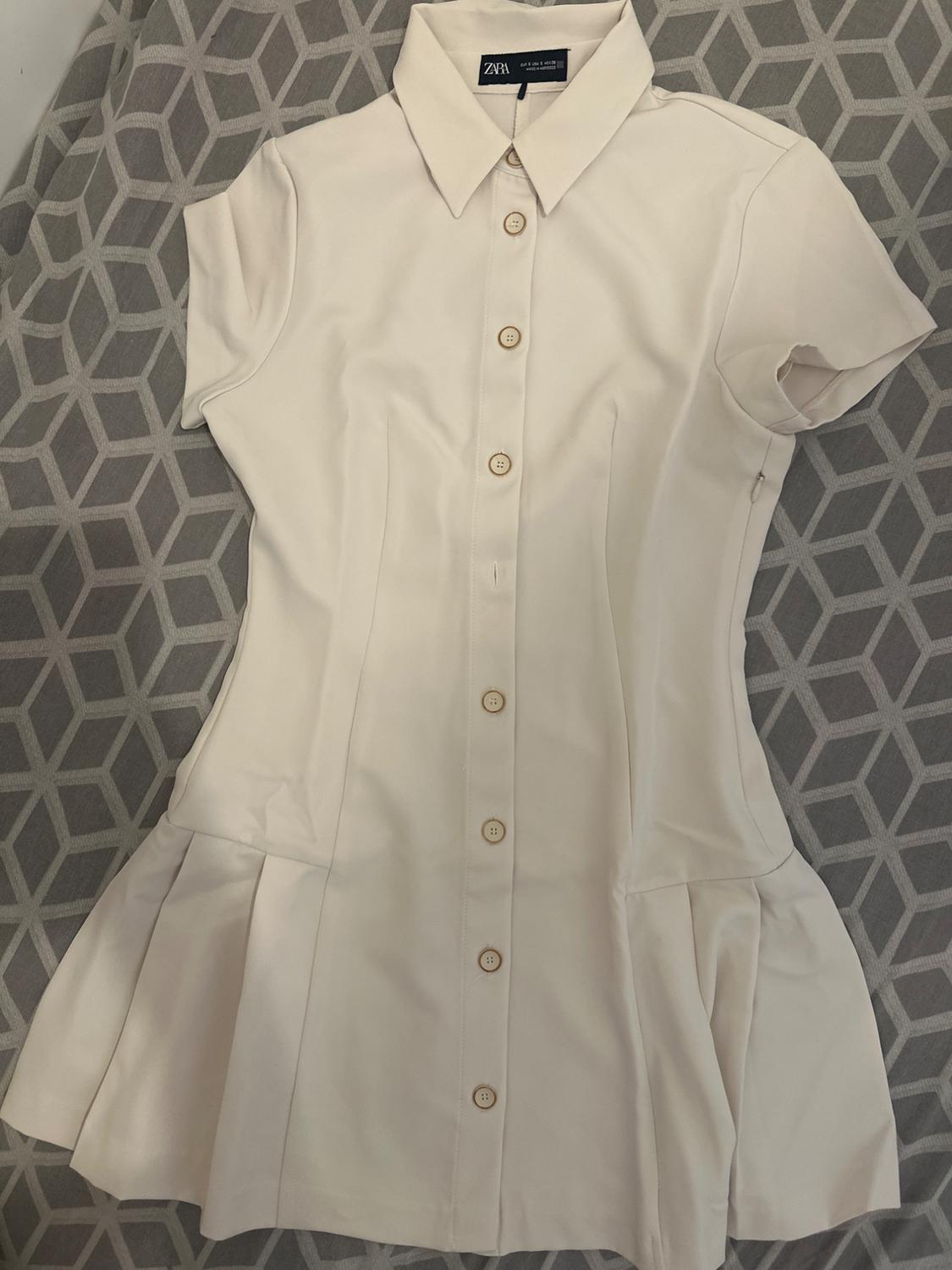 White short-sleeved dress with front buttons on a grey geometric pattern background. Visible label: Zara.