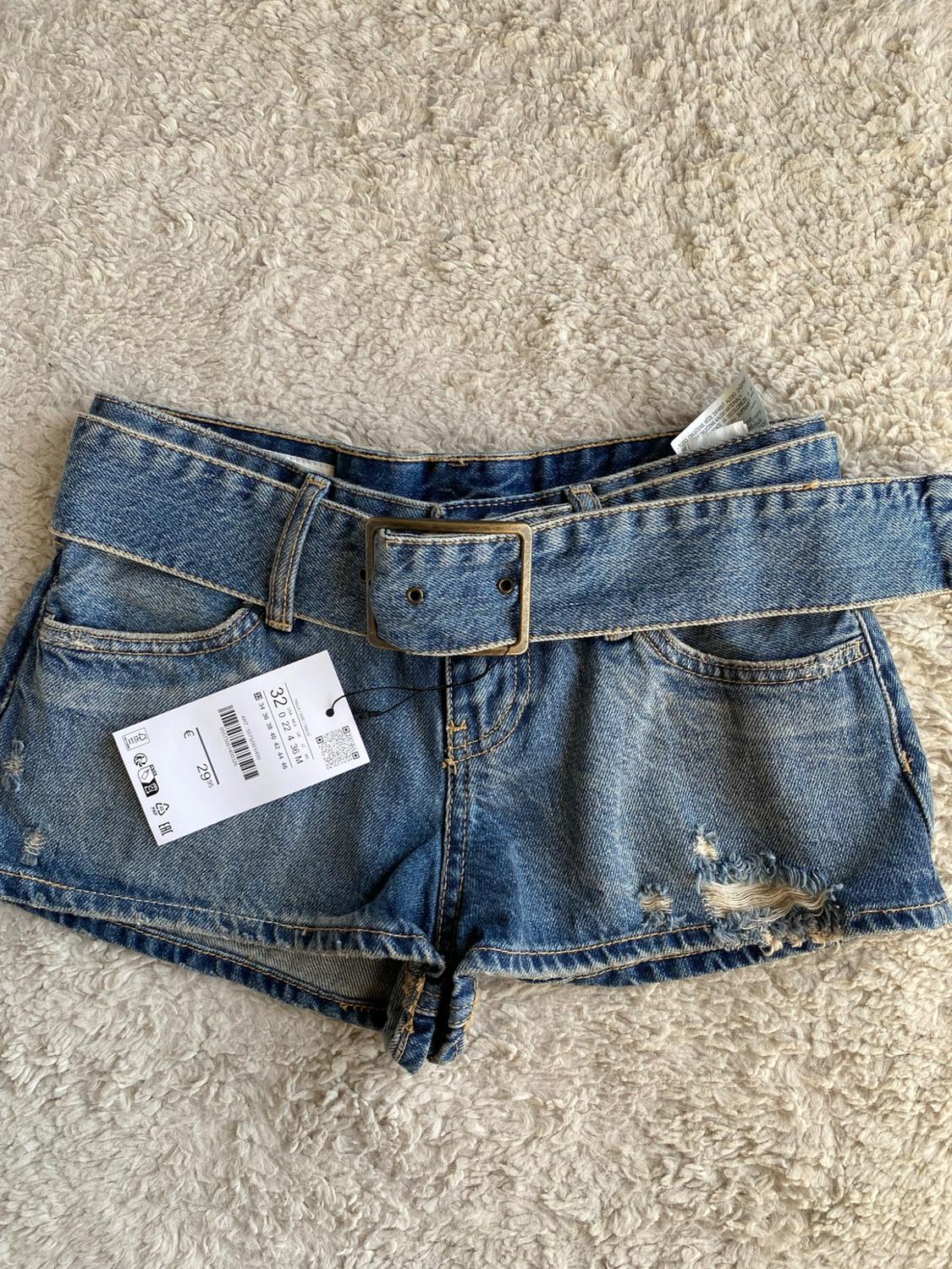 Blue denim shorts with a belt and rips on a plush carpet. Label with the text 32 and €29.95.
