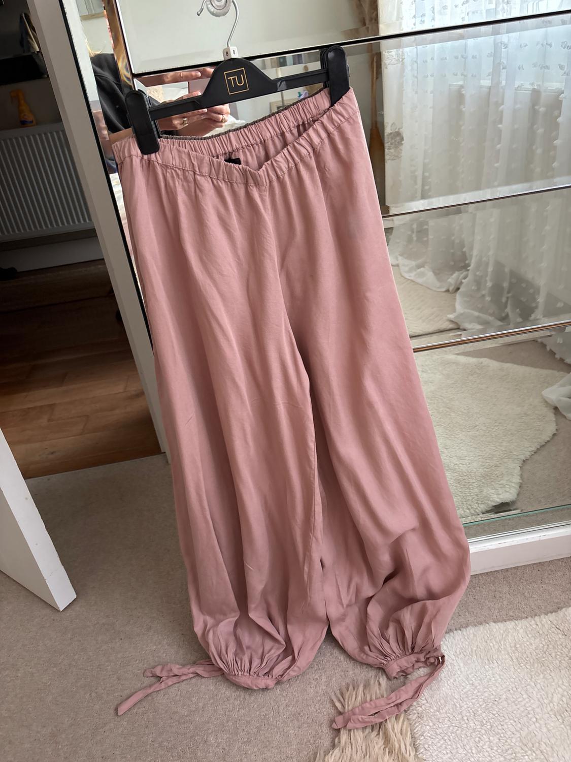 Pink trousers with an elasticated waist and ties at the ankles, hanging on a hanger with the text TU in front of a mirror in a room.