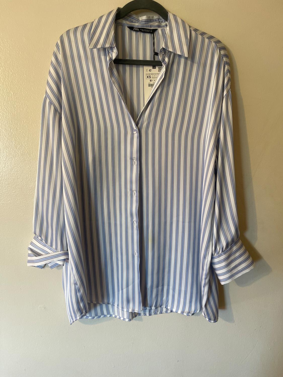 Blue and white striped shirt hanging on a black hanger. Visible tag with size XS. Beige wall background.