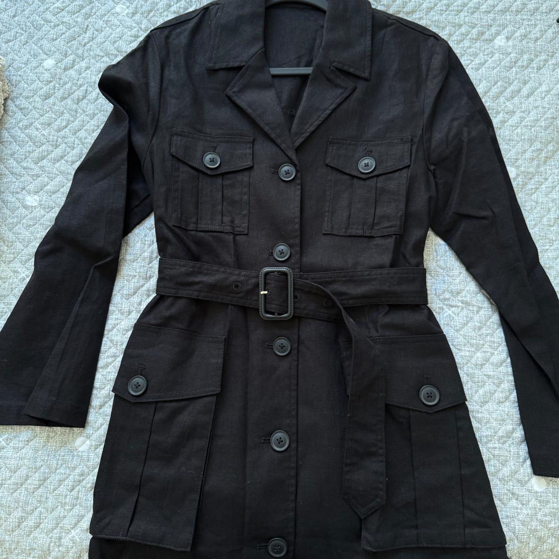 Black coat with belt and large buttons, four front pockets, over a gray quilt with a geometric pattern.