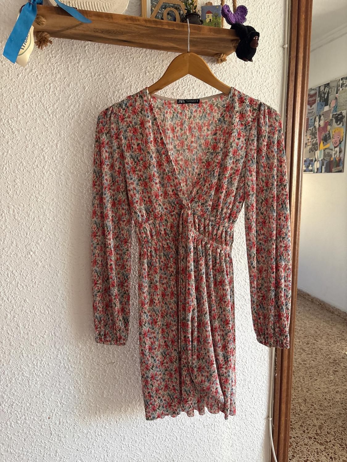 Short floral print ZARA dress with long sleeves, V-neck and gathered waist, hanging from a wooden hanger on a white wall.