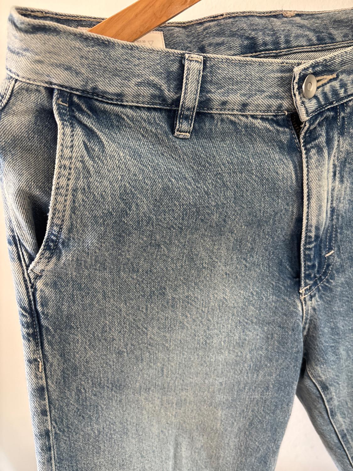 Close-up of light blue jeans on a wooden hanger. The texture, pocket, button, and an inner label with the text MEX 30 can be seen.