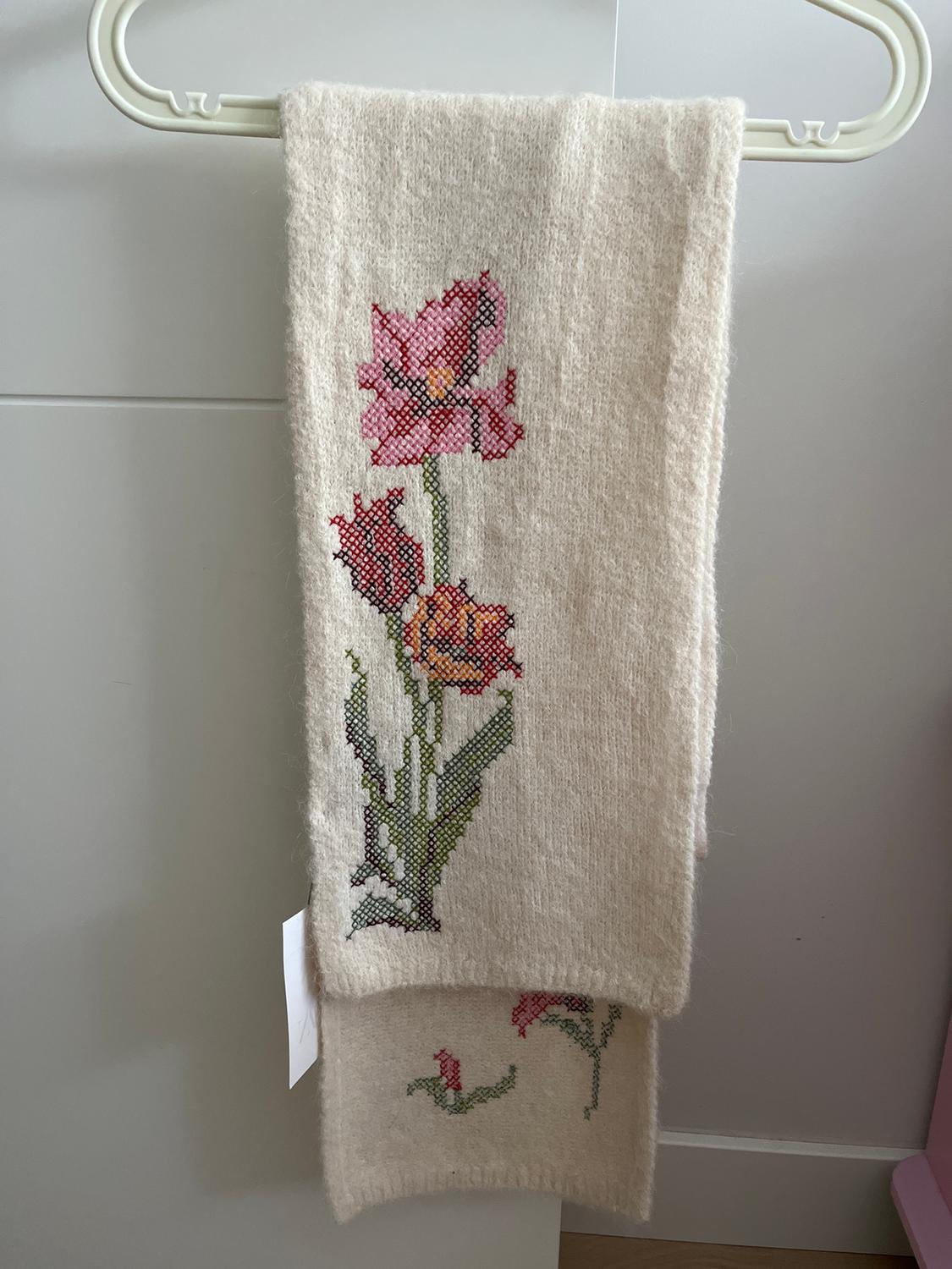 Beige scarf hanging on a white hanger, with embroidered floral design in pink and green tones. Light grey background.